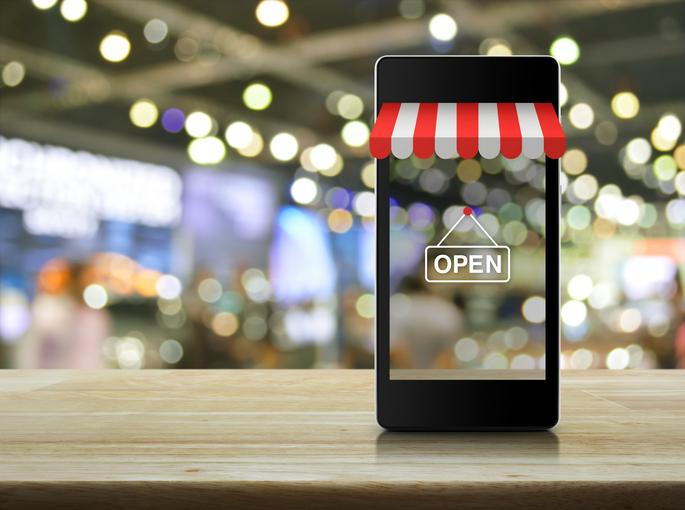 Modern smart mobile phone with on line shopping store graphic and open sign on wooden table over blur light and shadow of mall